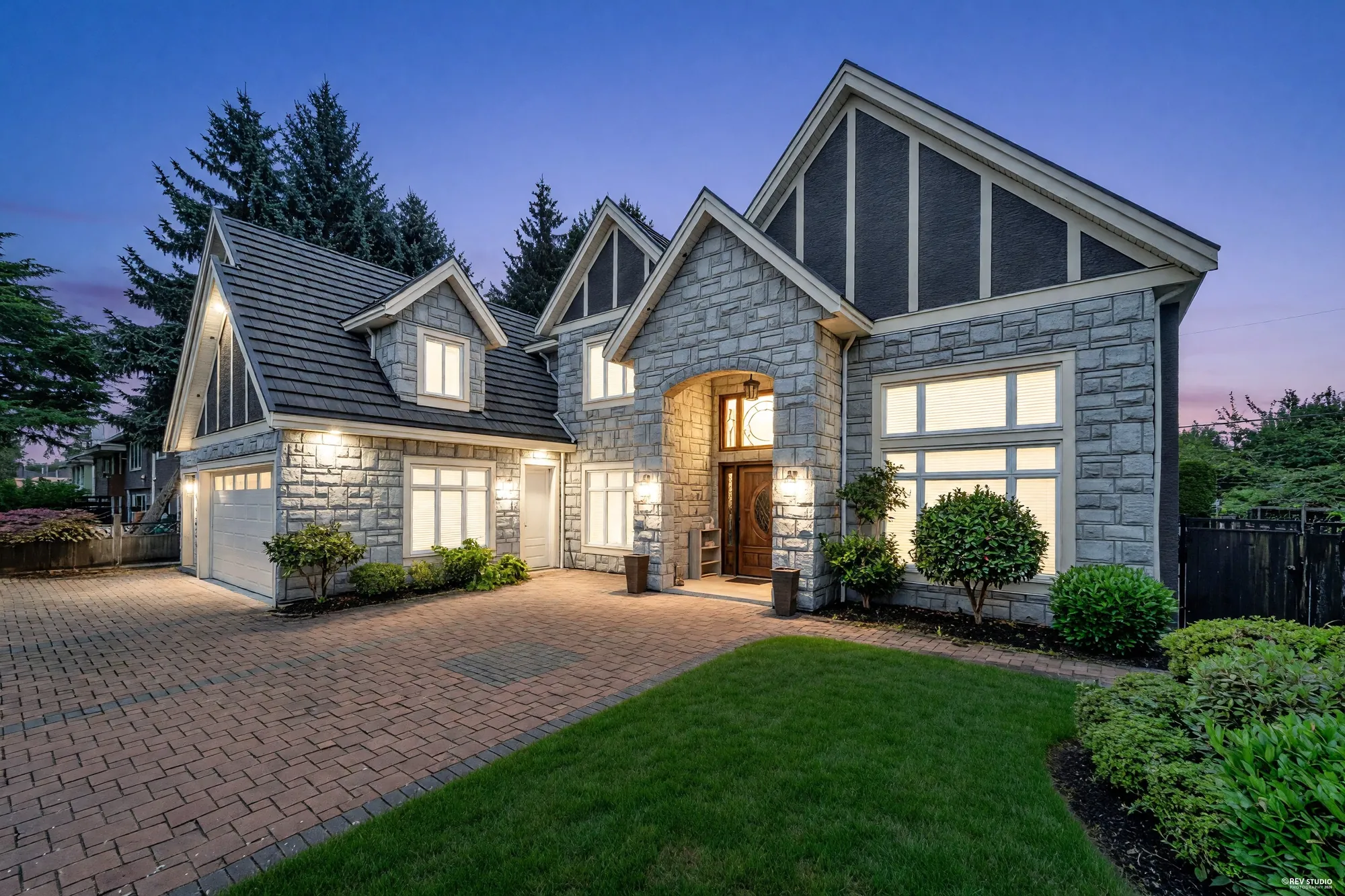 8080 Spires Road at dusk: the stone-fronted gables and the arched entry lit, the two garages and the brick drive in front