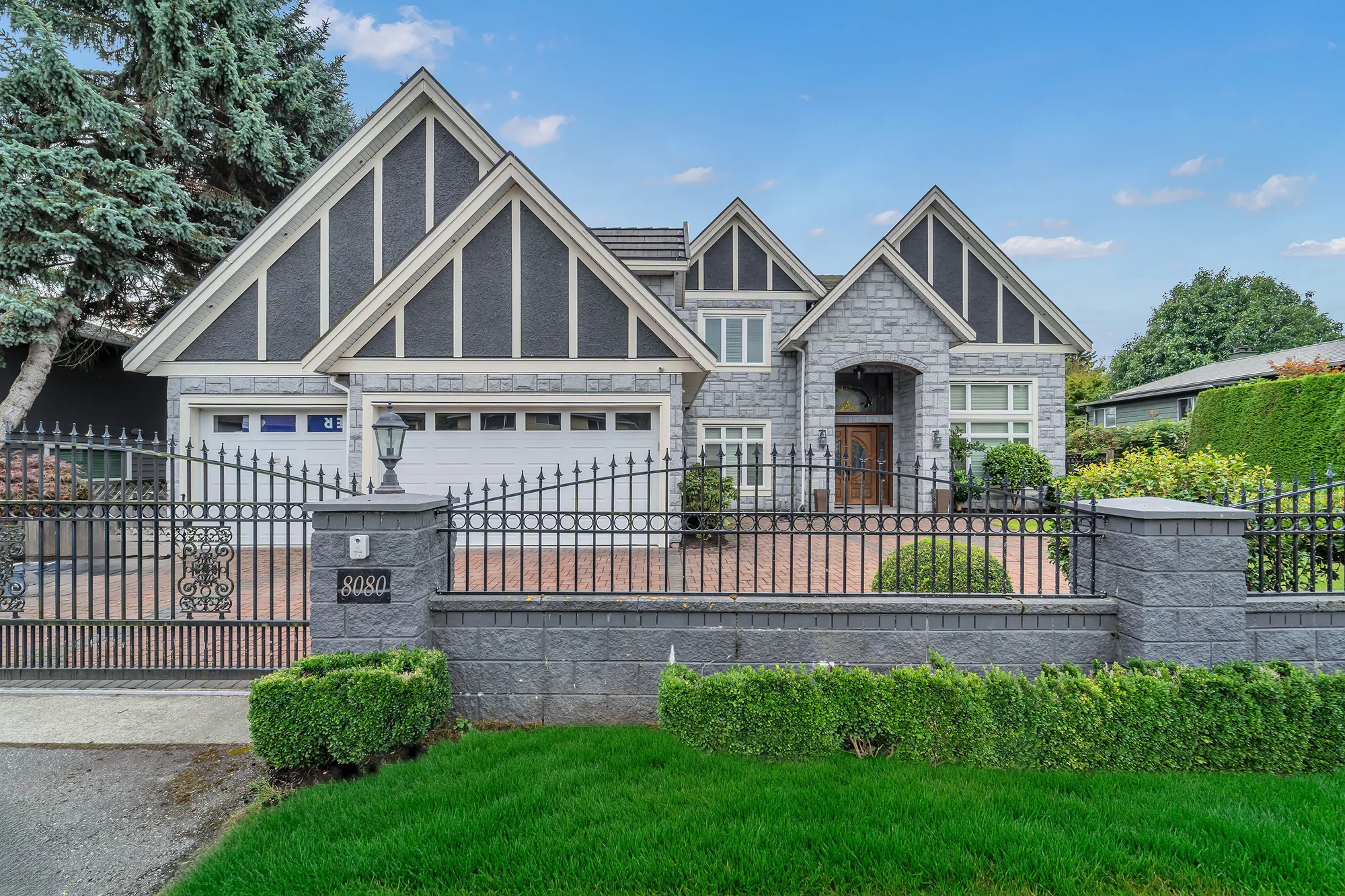 The house from the street in daylight: the iron fence and gate on a stone wall, the number on the pillar, the three gables behind