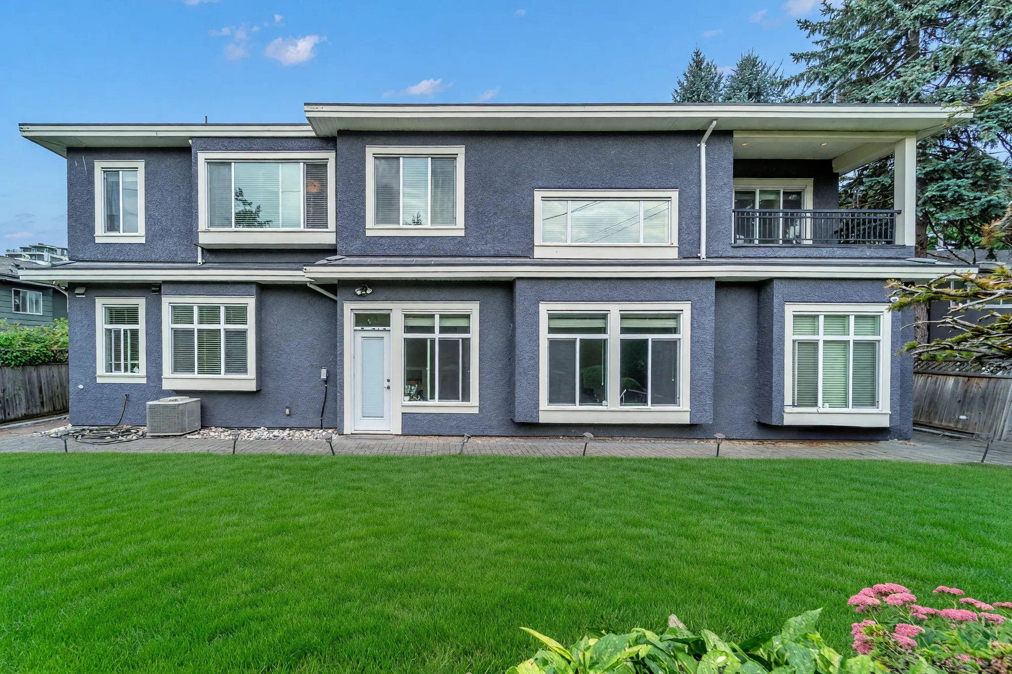 The back of the house in daylight: two storeys of grey stucco, the balcony at the upper right, the lawn in front